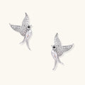 Pair of white gold-plated hummingbird stud earrings with pavé zirconia stones and teardrop crystal bodies on cream background