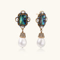 Ornate gold-plated earrings with iridescent abalone shell, crystals, blue stones, and white pearl drops on cream background.